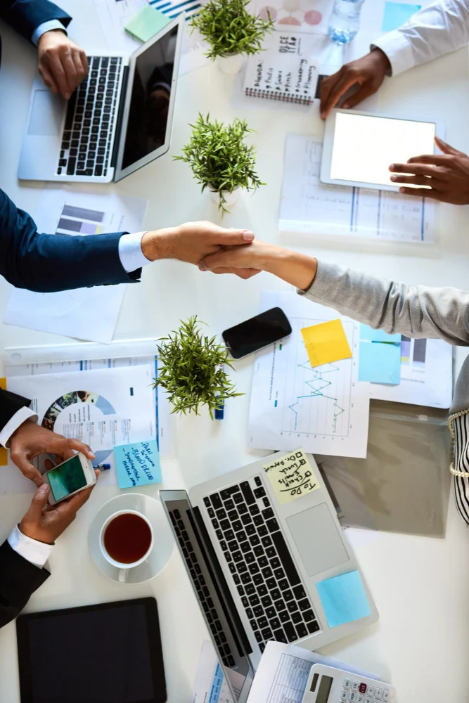 Business partners shaking hands over a desk of plans and reports